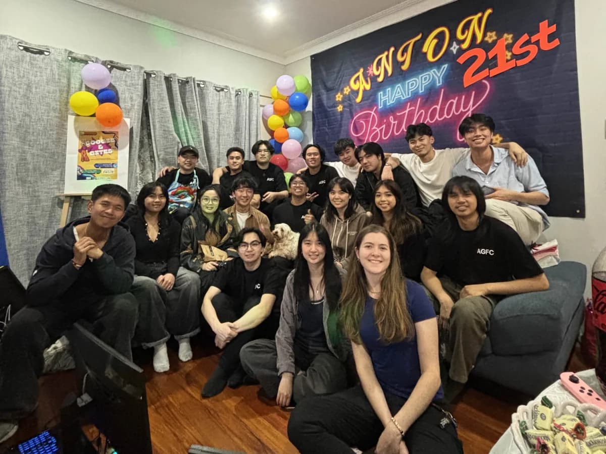 About twenty friends, several in AGFC shirts, crowded into a living room under a banner reading Anton Happy 21st Birthday
